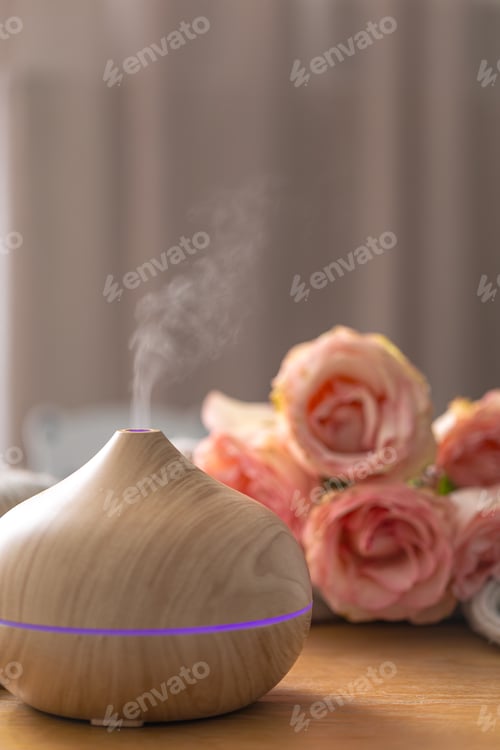 Preview: Close up wooden aroma diffuser releasing mist with roses background. Spa wellness device.