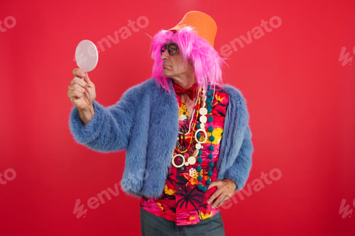 Preview: Senior man in funky outfit and pink wig checking reflection in a mirror