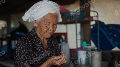 Preview: Grandmother wearing headscarf inspecting tiny item at home