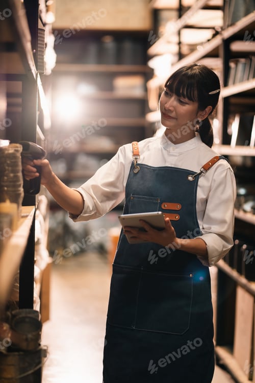 Preview: Woman in apron, warehouse aisle, checking stock, holding tablet