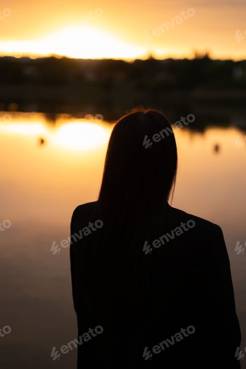 Preview: Backlit silhouette longhaired lonefigure at lakeshore golden sunset warm orange reflection peaceful