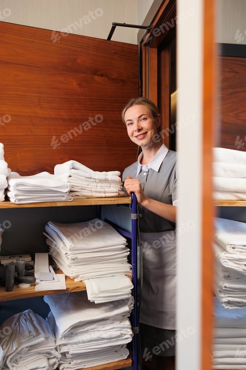 Preview: Friendly hotel maid standing with linen trolley and folded towels in hallway
