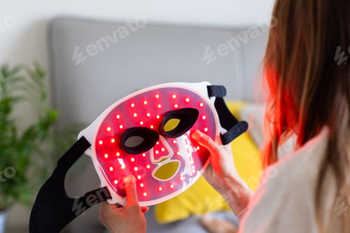 Preview: Woman holding LED light therapy mask at home