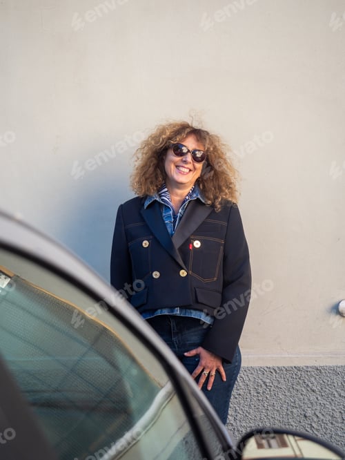 Preview: A Stylish Woman with Curly Hair Wearing a Blue Blazer and Sunglasses Standing Outdoors Next to a Car