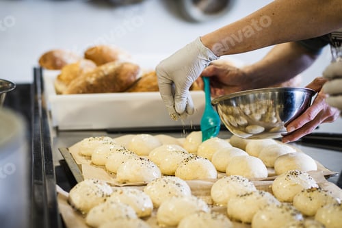 Preview: Baker Sprinkling Seasoning on Fresh Sourdough Bread Rolls Before Baking