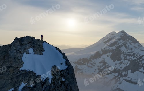 Preview: Solo Mountain Climber Standing on Rocky Snowy Peak at Sunrise Over Distant Alpine Ridges