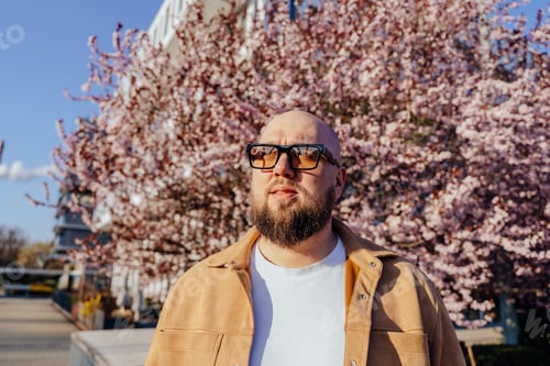 Preview: Serious bearded man in glasses looking forward among blooming trees