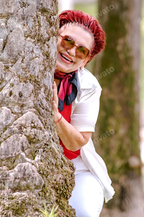 Preview: A stylish, smiling elderly woman in sunglasses playfully poses with a large tree