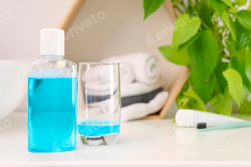 Preview: Bottle of mouthwash and glass on white table in bathroom, space for text