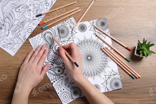 Preview: Woman coloring antistress picture with pencils at wooden table, top view
