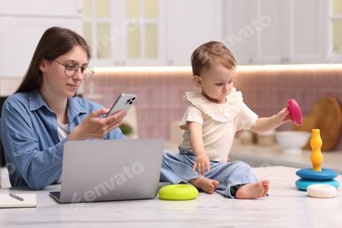 Preview: Young mother working and her baby playing with toy in kitchen at home