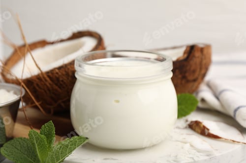 Preview: Delicious coconut yogurt in glass jar, halves of fruit, flakes and mint on table, closeup