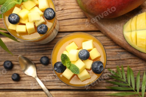 Preview: Delicious mango panna cotta with fruits and mint in glasses on wooden table, flat lay