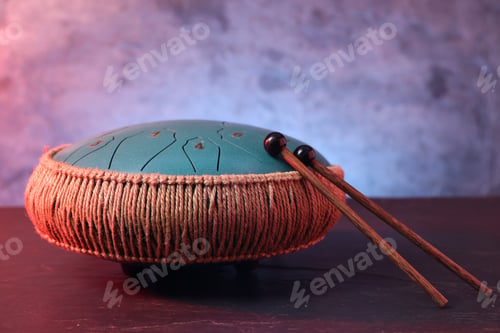 Preview: Steel tongue drum and mallets in red light on dark table textured wall, closeup