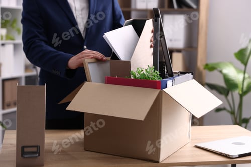 Preview: Dismissal. Man packing his personal belongings into cardboard box at wooden table in office, closeup