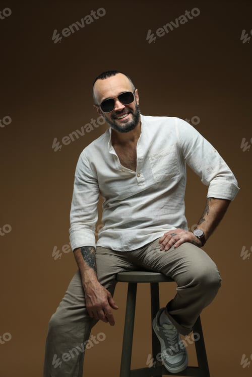 Preview: Fashion model. Handsome man in sunglasses on stool against brown background