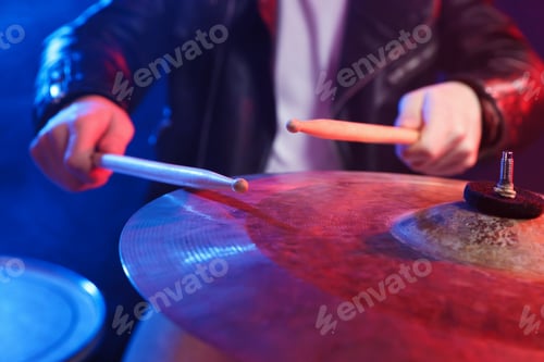 Preview: Man with drumsticks playing cymbal in color lights against dark background, closeup