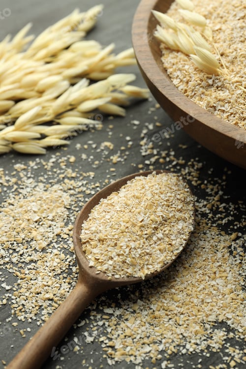 Preview: Oat bran in bowl, spoon and spikes on black table, closeup