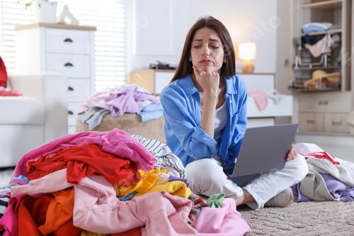 Preview: Woman with laptop surrounded by different messy clothes indoors