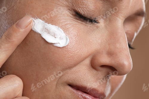 Preview: Beautiful woman applying cream onto her face on beige background, closeup