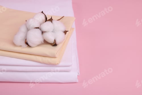 Preview: Stack of fresh bed linens and cotton flowers on pink background, closeup. Space for text