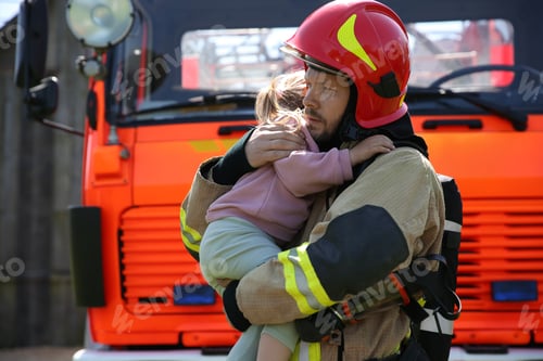 Preview: Firefighter in uniform holding rescued little girl near fire truck outdoors. Save life