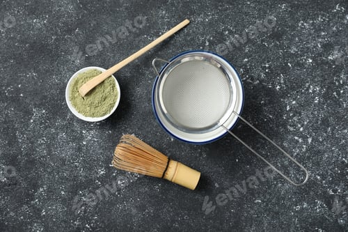 Preview: Green matcha powder, scoop, bamboo whisk, sifter and ceramic bowl on black textured table, flat lay