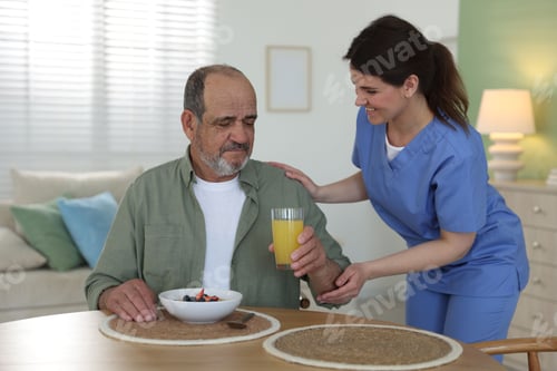 Preview: Caregiver giving breakfast to elderly man at wooden table indoors