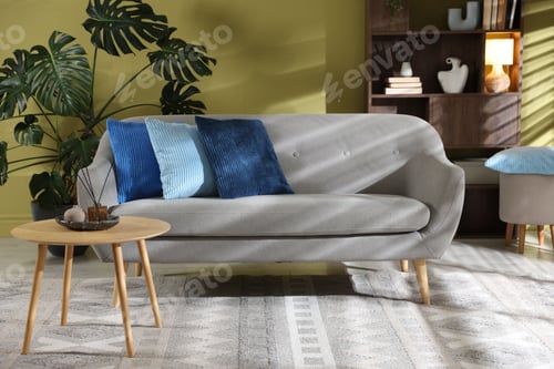 Preview: Soft pillows on grey sofa, side table, shelving unit and houseplant near green wall indoors