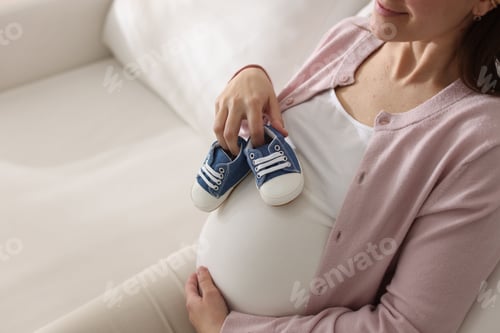 Preview: Pregnant woman with baby shoes sitting on sofa, closeup. Space for text