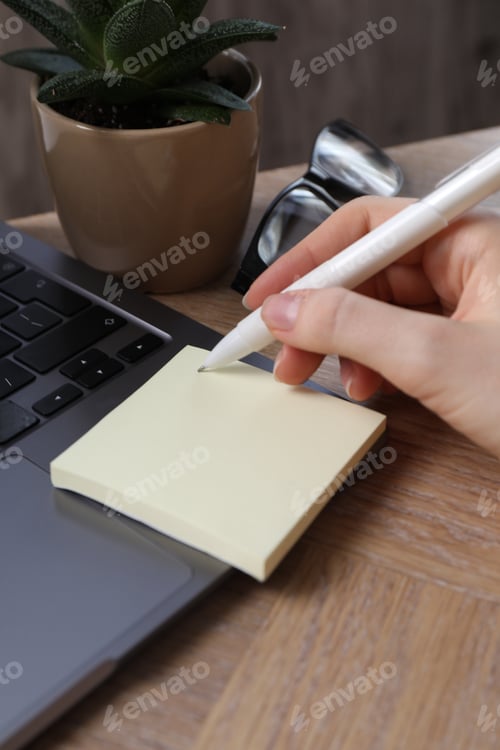 Preview: Reminder. Woman writing on sticky note at wooden table, closeup
