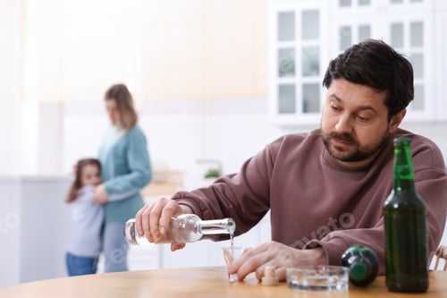 Preview: Alcohol addiction. Man drinking vodka at wooden table while his scared wife and daughter standing in
