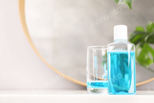 Preview: Bottle of mouthwash and glass on white table in bathroom, space for text