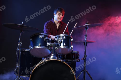 Preview: Smiling teenage boy with drumsticks learning how to play drums in color lights and smoke against dar