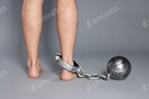 Preview: Man with metal ball and chain on his leg against grey background, closeup