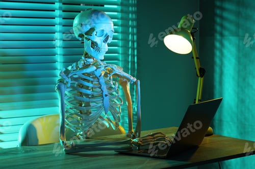 Preview: Waiting concept. Human skeleton sitting at wooden table with laptop indoors