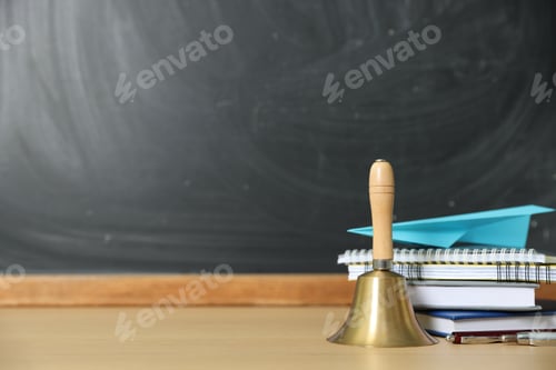 Preview: School holidays. Golden bell, paper plane, notebooks, books and pens on wooden table near chalkboard