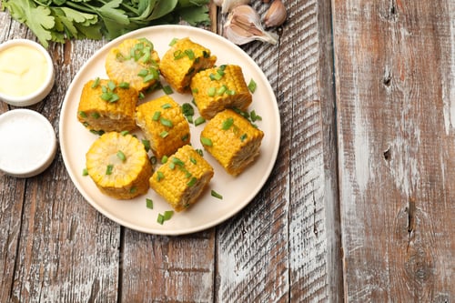 Preview: Pieces of boiled corn cobs with green onion served on wooden table, flat lay. Space for text