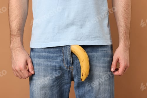 Preview: Urology. Man with banana in his pants on beige background, closeup