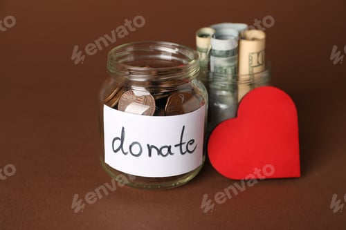 Preview: Donation jars with money and red decorative heart on brown background, closeup