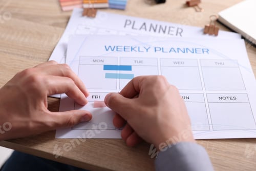 Preview: Man using weekly planner at wooden desk indoors, closeup