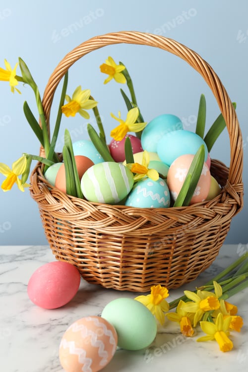 Preview: Painted Easter eggs and beautiful daffodils in wicker basket on white marble table against light blu