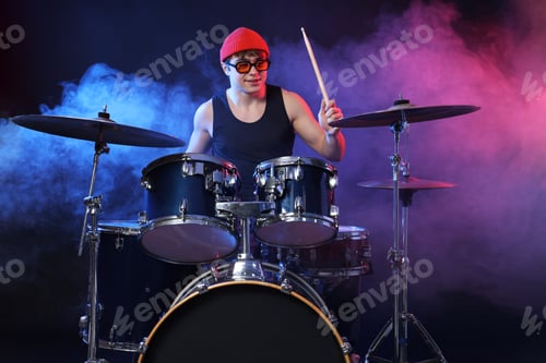 Preview: Smiling teenage boy in glasses and hat learning how to play drums in color lights and smoke against