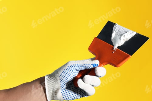 Preview: Handyman holding putty knife with plaster on yellow background, closeup. Space for text