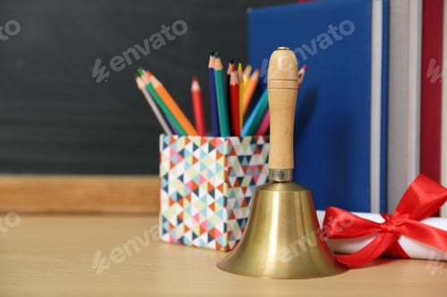 Preview: School holidays. Golden bell, diploma, books and pencils on wooden table near chalkboard, closeup. S