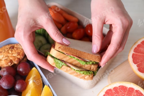 Preview: Woman putting delicious sandwich into lunch box at light wooden table, above view