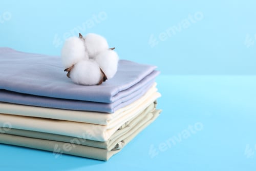 Preview: Stack of fresh bed linens and cotton flower on light blue background, closeup. Space for text