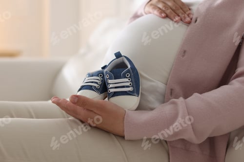 Preview: Pregnant woman with baby shoes sitting on sofa at home, closeup