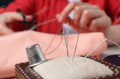 Preview: Woman cutting sewing thread over cloth, focus on pin cushion with needles and thimble