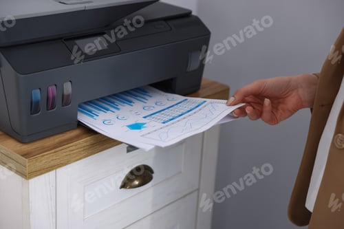Preview: Woman taking printed sheet of paper from printer at chest of drawers indoors, closeup